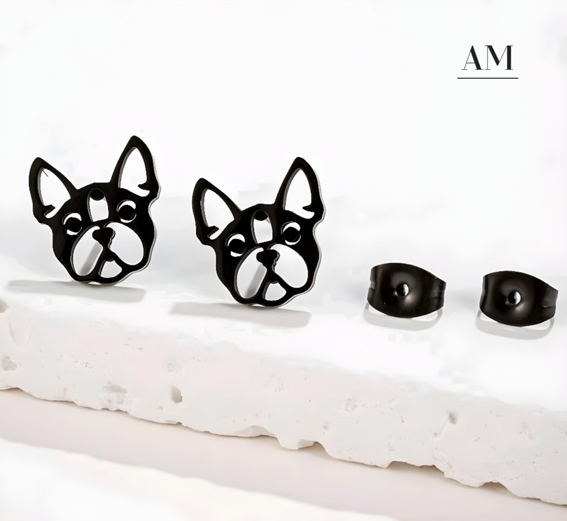 A pair of black stainless steel bulldog earrings displayed on a stand, with the brand name 'AM' visible at the top.