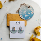 Earring set on a card with 'AM' branding, surrounded by yellow flowers and a decorative coaster.