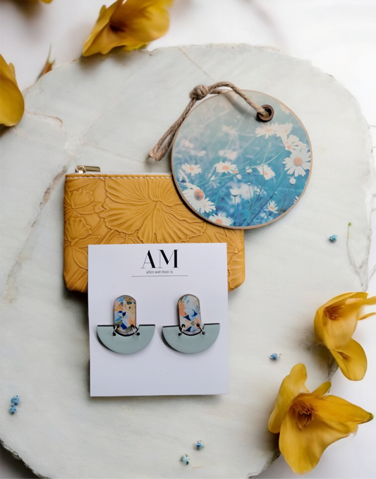 Earring set on a card with 'AM' branding, surrounded by yellow flowers and a decorative coaster.