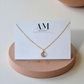 Gold necklace on a card with 'AM' branding on a beige stand.